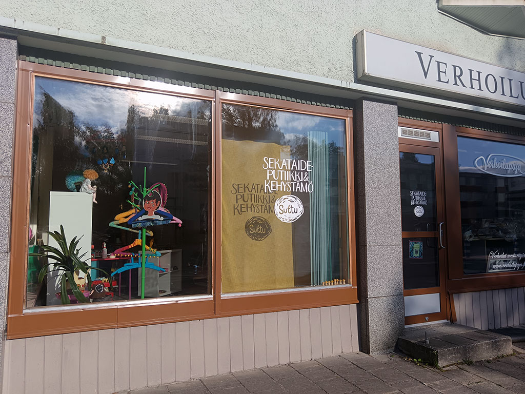 Kaupan ikkunoissa värikkäitä koristeita ja tekstiä, ulkopuolella harmaa julkisivu ja puinen sisäänkäynti.